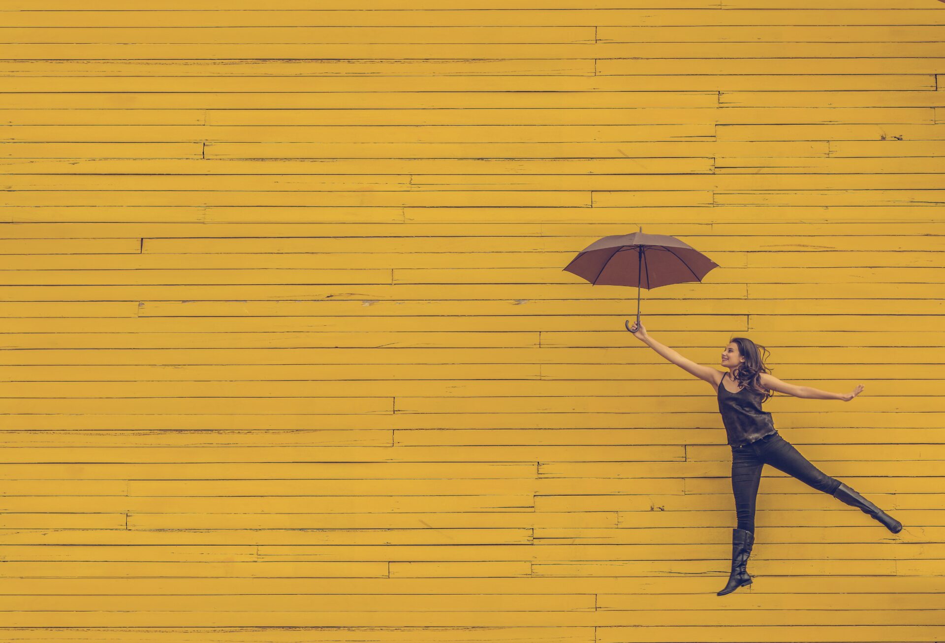 Eine junge Frau vor gelben Brettern hält einen braunen Regenschirm in die Höhe.