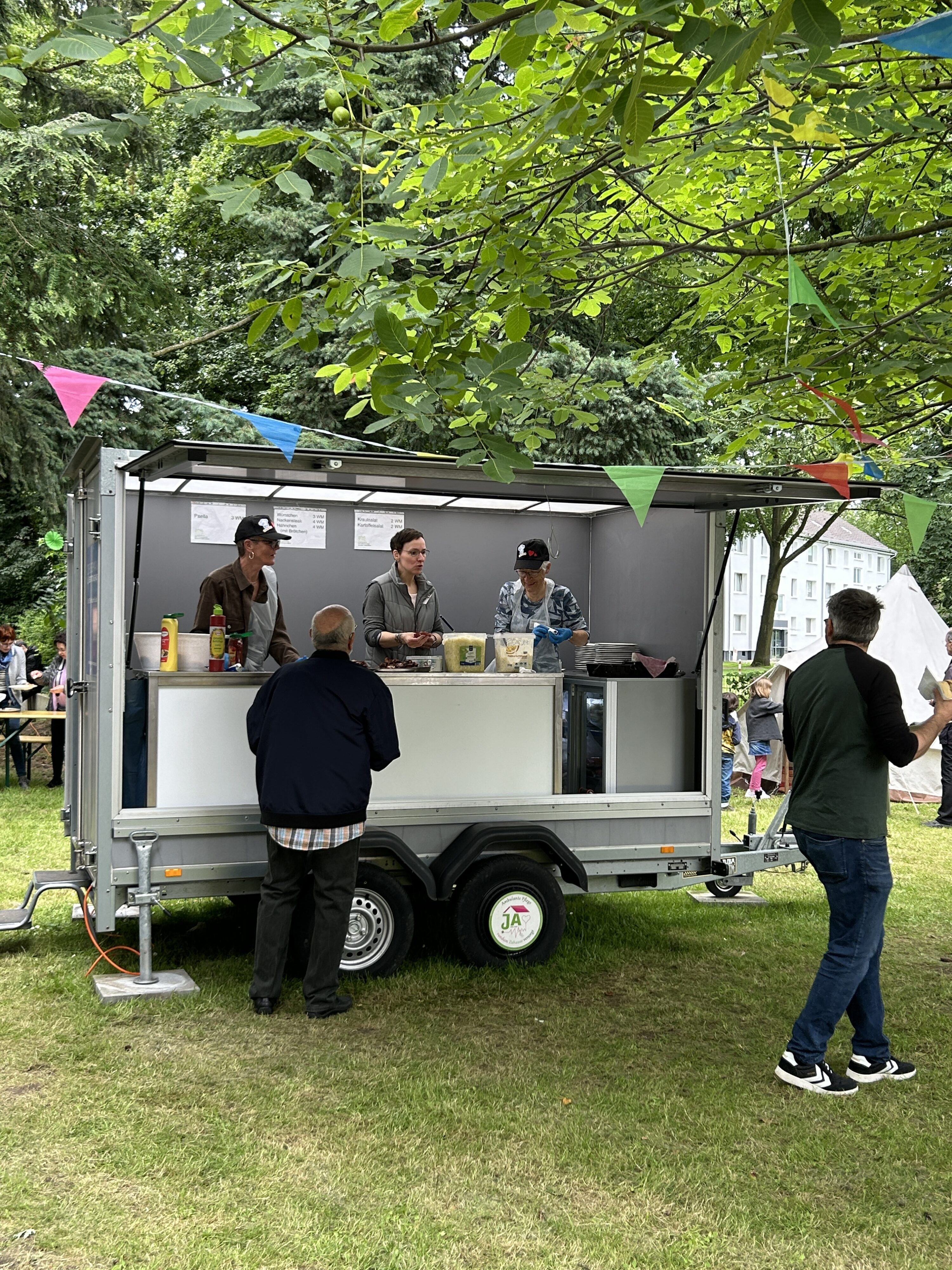 Im Garten des Martin-Niemöller-Hauses steht ein Festwagen, auf dem drei Personen Essen ausgeben. Im Vordergrund zwei Personen mit Essen in der Hand.